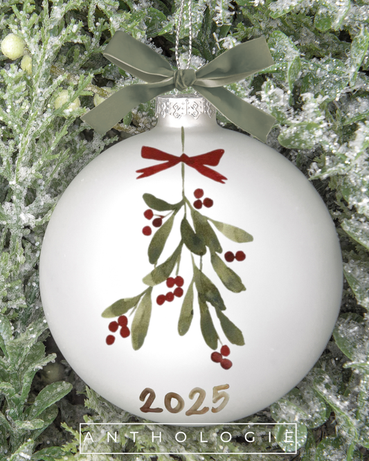 White Christmas ornament with mistletoe design and '2025' on a green leafy background, featuring the brand 'Anthologie'.