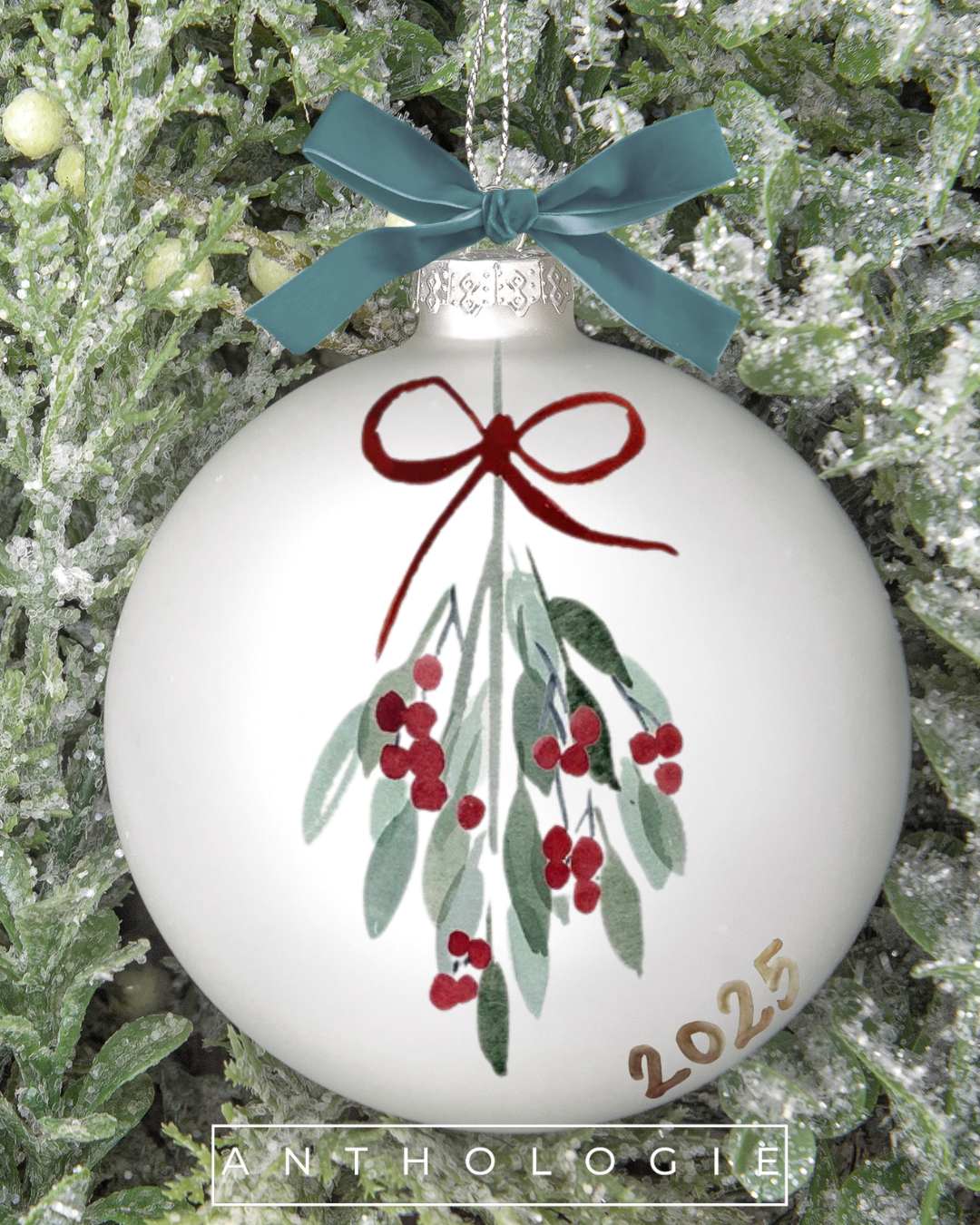 White Christmas ornament with mistletoe design and blue ribbon on a tree, featuring the brand 'Anthologie'.